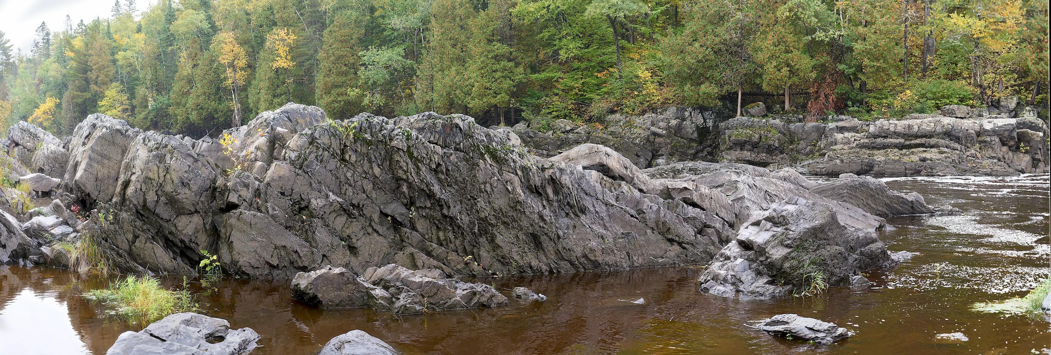 A rock formation in a still portion of a wide river. Trees are in the background.